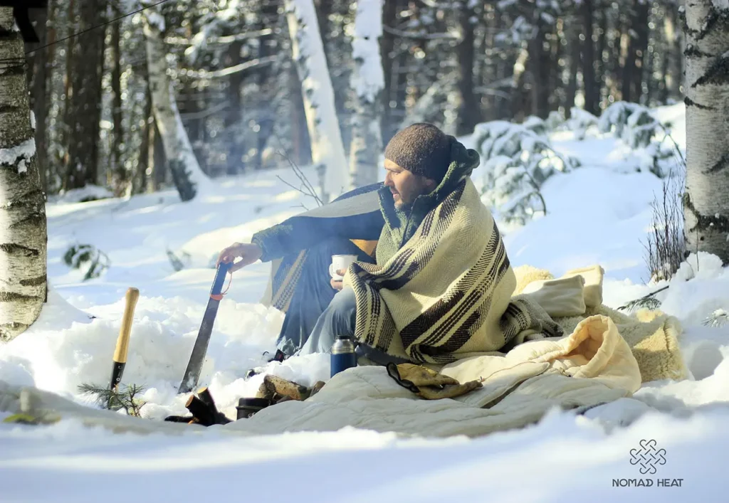 Campingman i snön sittande på en ullmatta täckt med en ullfilt bredvid en ullsovsäck.