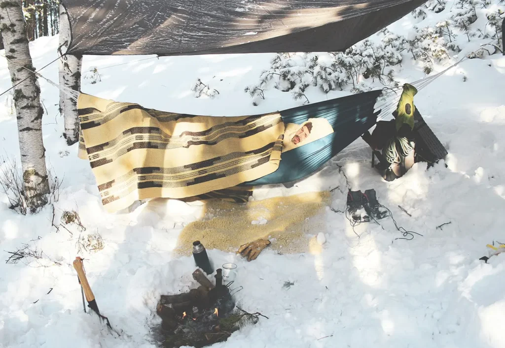 man som campar i snön i en hängmatta i en ullsovsäck täckt med en filt