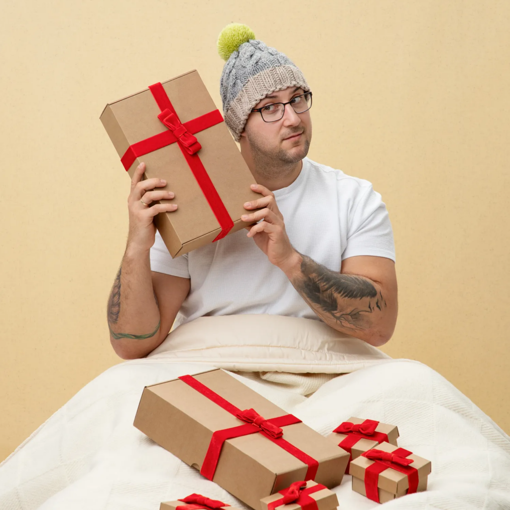 man in white t-shirt and winter hat holding a christmas present next to his face